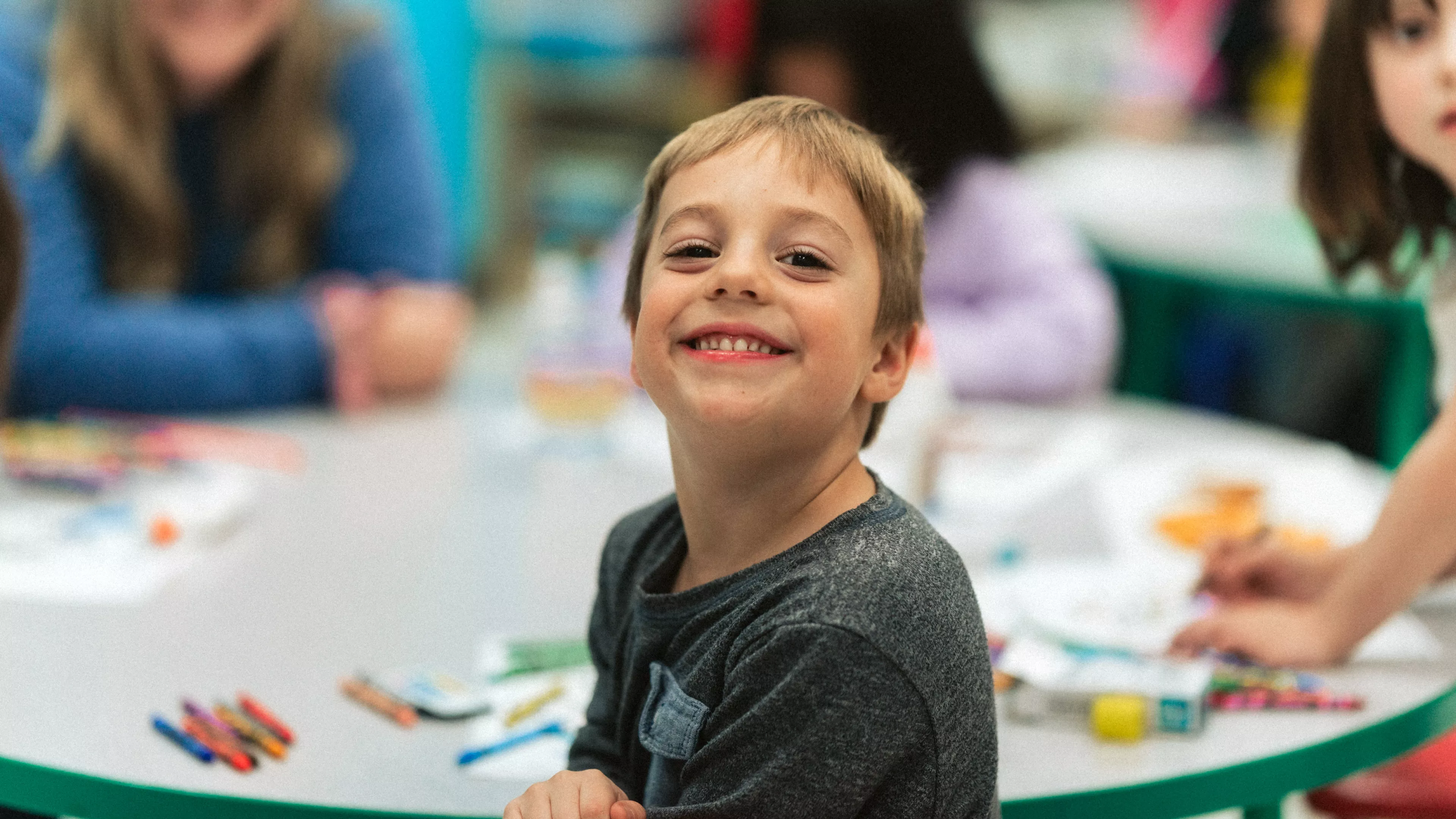 A young child smiles while sitting at a table with a smiling teacher in the background.