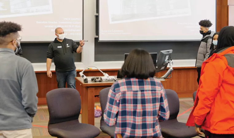 A teacher stands at the front of a classroom addressing students of college age. They are all wearing protective masks as they circle the teacher listening to his speech.